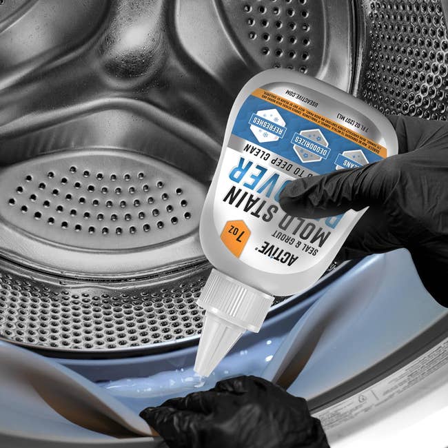 Gloved hands clasp  a mold and stain remover bottle, pouring it into an unfastened  washing machine
