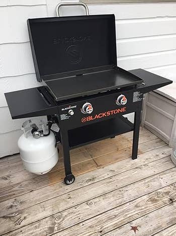 Outdoor Blackstone state griddle with 2 burners connected a woody deck, featuring broadside shelves and a propane tank. Suitable for cooking and grilling