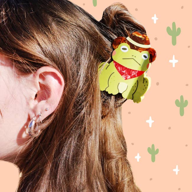 Close-up of a person wearing a frog-themed hair accessory with cowboy hat details. Cactus illustrations decorate the background