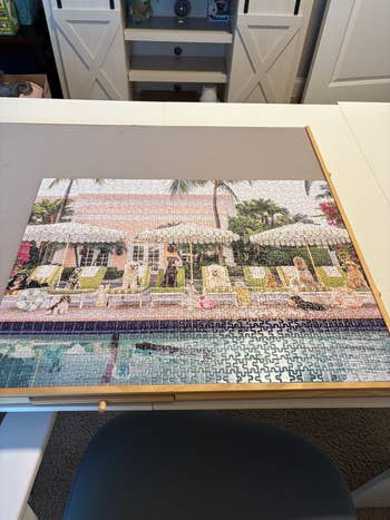 Finished jigsaw puzzle depicting a luxury poolside scene with sun loungers and umbrellas on a table in a home setting