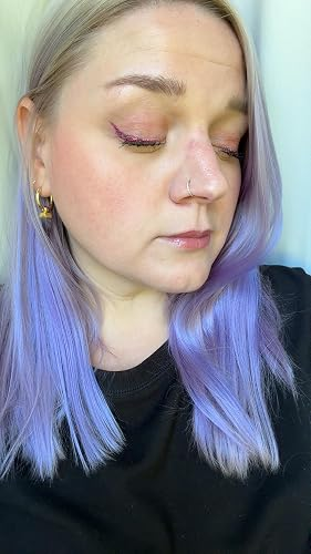 Person with lavender hair and nose ring, wearing a black top, showcasing shimmery eye makeup and gold hoop earrings