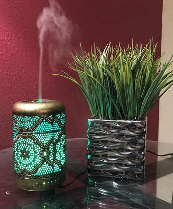 another reviewer's gold metal vintage diffuser actively working with green lights on and emitting vapor into the air, sitting next to a plant