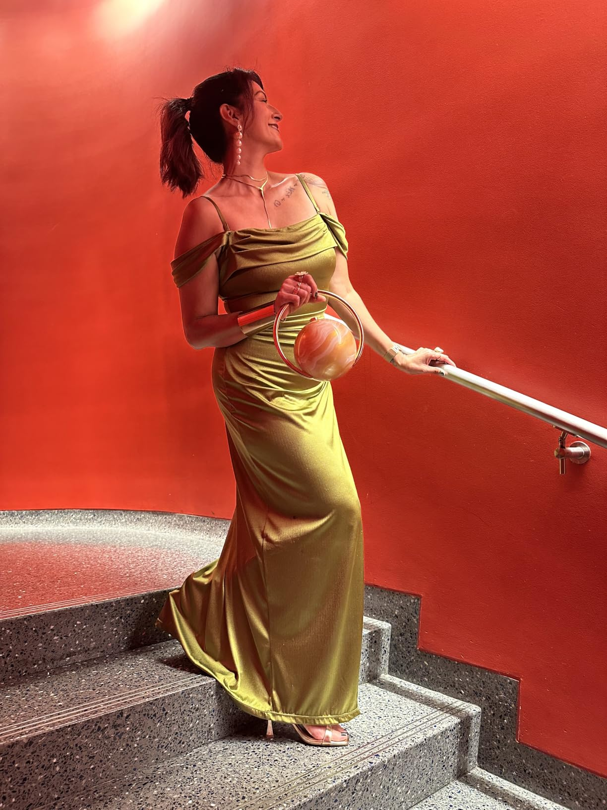Person successful  elegant, off-shoulder silk gown holding a stylish, circular  handbag connected  stairs with reddish  backdrop