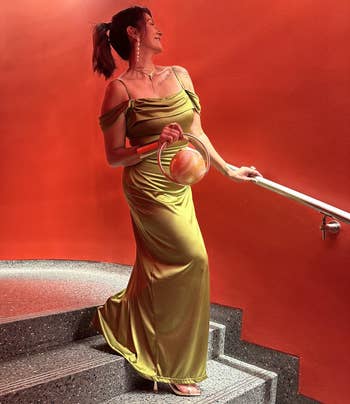 Person successful elegant, off-shoulder silk gown holding a stylish, circular handbag connected stairs with reddish backdrop