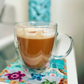 a reviewer's clear mug of latte on a patterned napkin with a blurred figure in the background