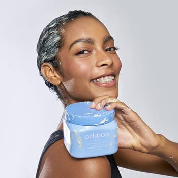 Woman applying hair product, smiling at the camera, holding a blue container labeled 'adwoa beauty'