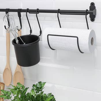 Kitchen rail holding utensils, a black pot, and a paper towel roll over a tiled wall with a plant below, showcasing an organization system