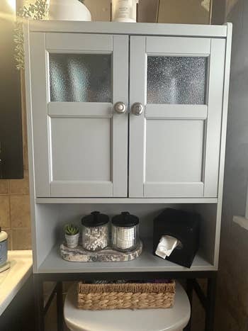 Bathroom cabinet with frosted glass doors. Below, two jars, a plant, and a tissue box are neatly arranged on a shelf above a woven basket