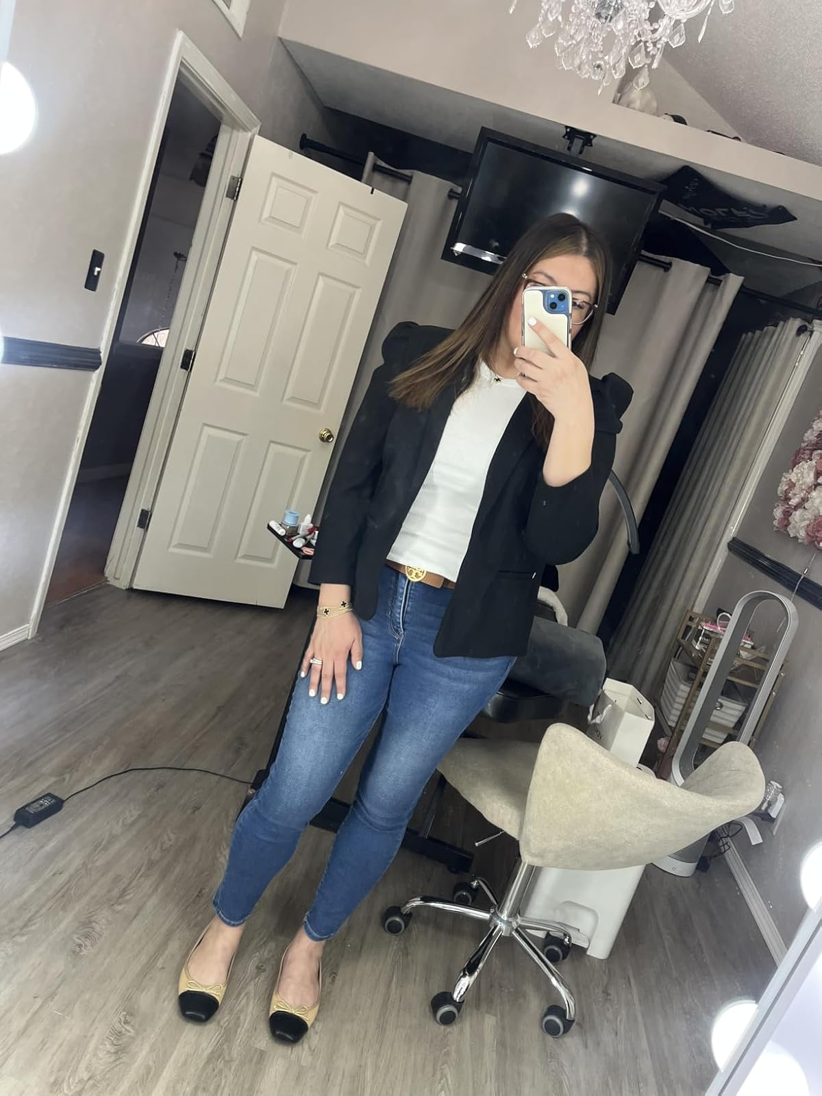 Person taking a mirror selfie in a salon, wearing a black jacket, white top, blue jeans, and flats, with styling chairs and tools in the background