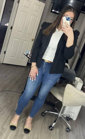 Person taking a mirror selfie in a salon, wearing a black jacket, white top, blue jeans, and flats, with styling chairs and tools in the background