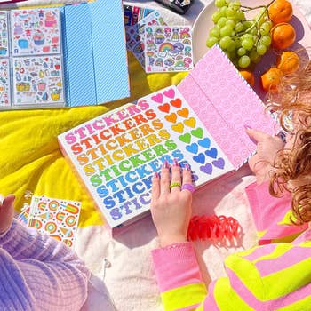 Two people are sitting at a table covered in colorful stickers, small sticker books, grapes, and oranges, browsing through a sticker book