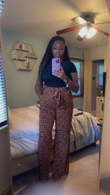 Person taking a mirror selfie in a bedroom, wearing a black top and pattern pants, standing confidently