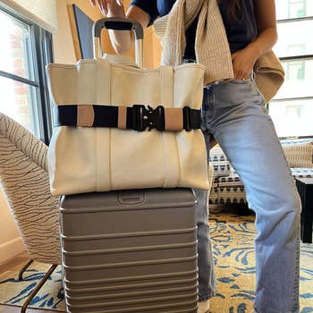 A person wearing a beige scarf over a blue top and jeans stands beside a beige tote bag secured on a silver suitcase in a stylish hotel room