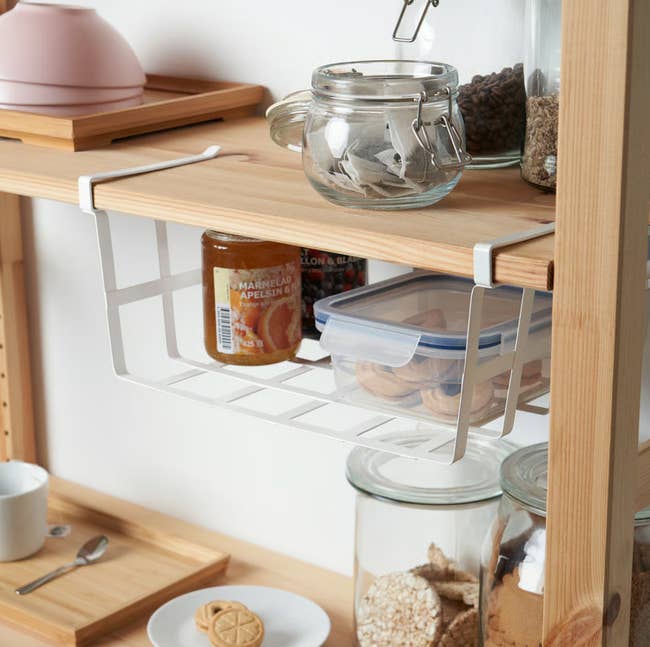 Undershelf basket with jars, cookies, and kitchen items on a wooden shelf, demonstrating space-saving storage for home organization