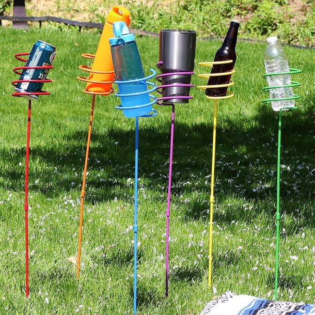 Five garden stakes with spiral holders containing different types of drinkware in a grassy yard