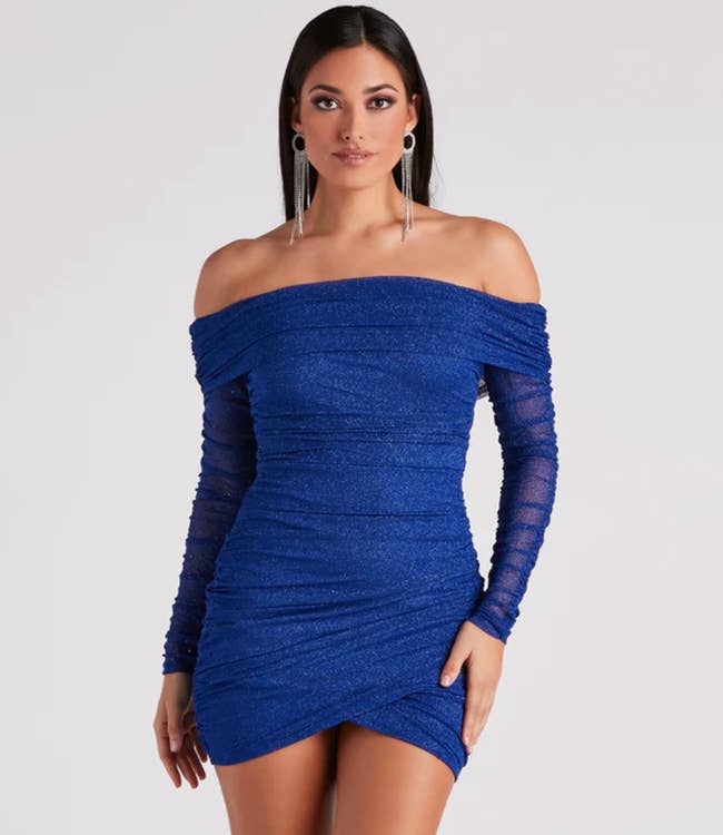 Model is wearing a bold blue glittery off shoulder bodycon dress with mesh long sleeves