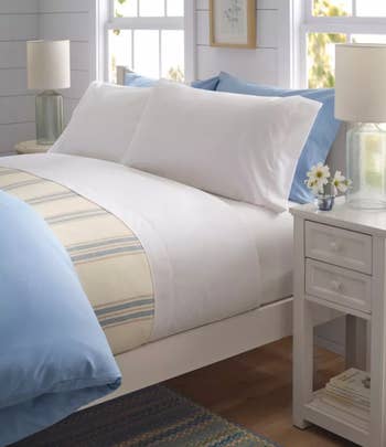 Bedroom scene featuring a neatly made bed with white bedding, blue accents, and a patterned throw blanket; a bedside table with a lamp and flowers