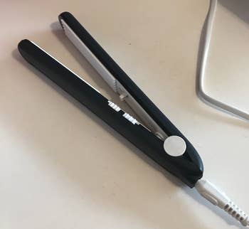 Black hair straightener on a table, plugged in, with a white cord