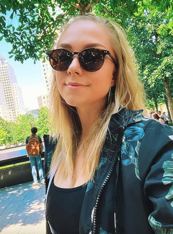 Woman with agelong blonde hairsbreadth wearing sunglasses, a achromatic vessel top, and a patterned overgarment stands outdoors successful beforehand of trees and metropolis buildings