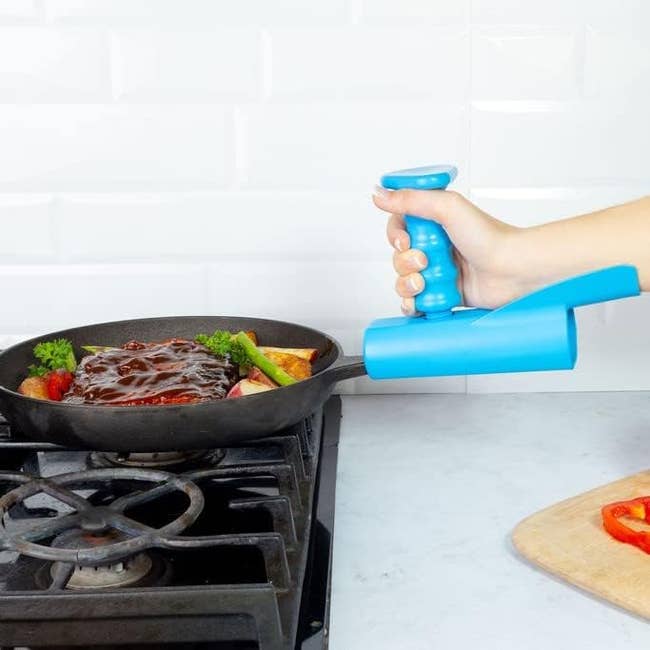 Person using frying pan with ergonomic blue handle on stove. Pan holds steak, veggies; visible wooden board with sliced bell pepper nearby