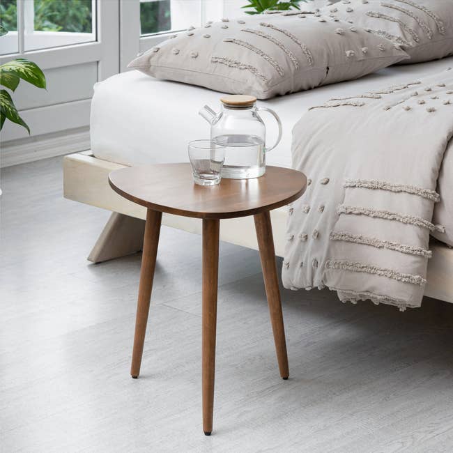 A wooden bedside table with a glass water pitcher and glass. A modern bed with textured bedding is in the background