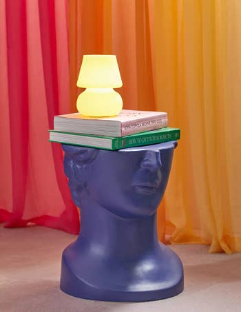 blue sculptural bust styled as a table with a stack of books and a small lamp on top