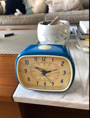 blue rectangular analog alarm clock with rounded edges perched on a table 