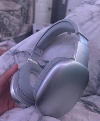 A close-up of someone's hand holding silver over-ear headphones while on a bed with a patterned blanket in the background