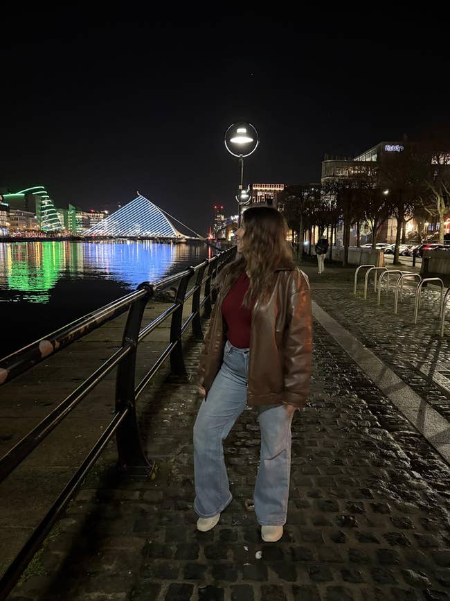 A person in a leather jacket and jeans stands on a cobblestone path by a waterfront, illuminated cityscape in the background at night