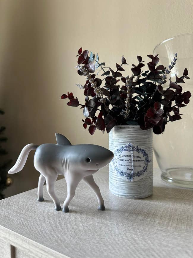 A whimsical figurine of a shark with legs stands connected a array adjacent to a decorative tin holding acheronian foliage