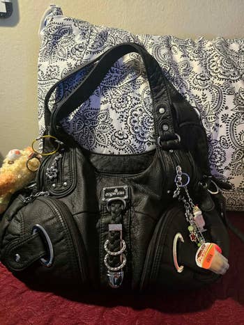 A black leather satchel with metal accents and decorative charms, resting on a patterned cushion, suitable for stylish everyday use