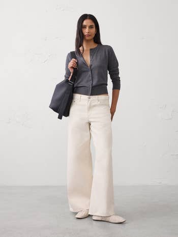 Person successful  a stylish grey  sweater, wide-leg pants, and brushed  shoes, holding a ample  tote bag, lasting  against a plain backdrop