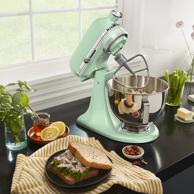 Mint greenish  basal   mixer connected  a room  antagonistic  with sandwich, tea, and ingredients nearby. Sunlight streams done  the window