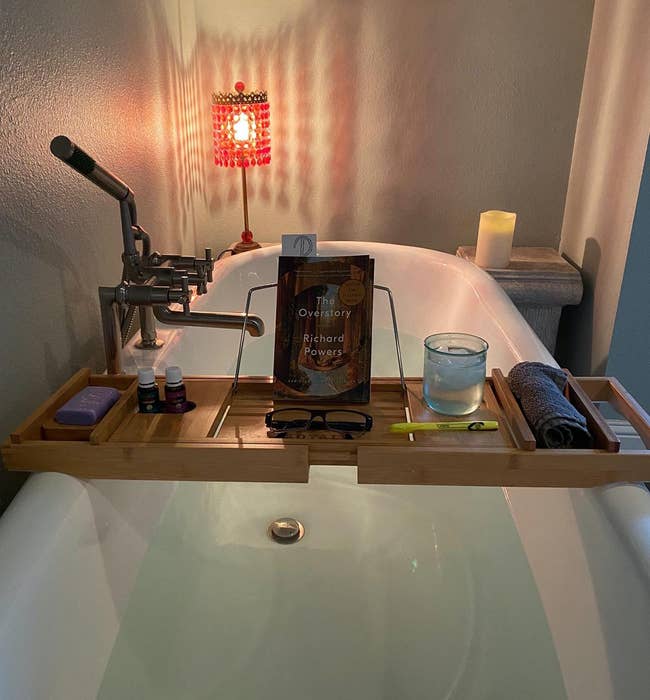 Bath caddy over tub with book, glasses, candle, personal care items, and a rolled towel