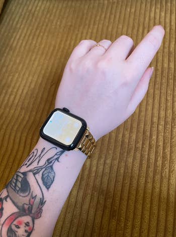 Person's wrist with a smartwatch featuring a gold band and several tattoos visible on the arm