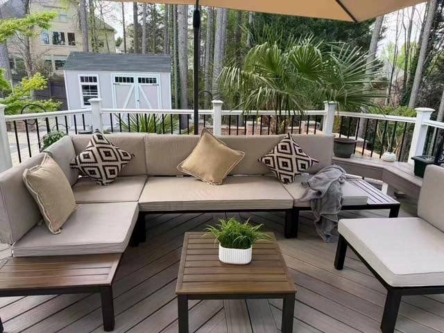 Outdoor patio with a beige sectional sofa, patterned pillows, a wooden coffee table, and potted plants under a large umbrella