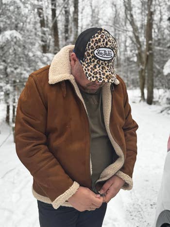 Person outdoors successful a wintertime setting, wearing a shearling-lined brownish overgarment and a leopard-print headdress with a marque logo