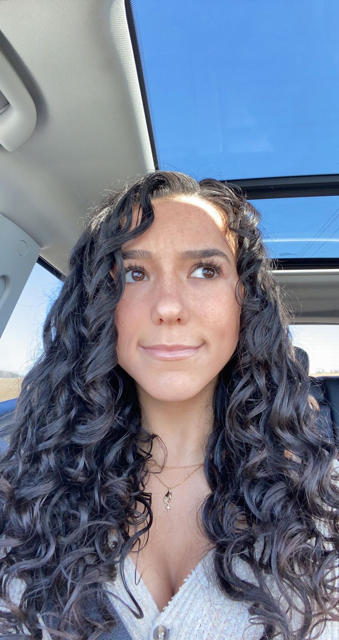 Person with long, curly hair sits in a car with a sunroof. They wear a textured top with a delicate necklace, gazing upward