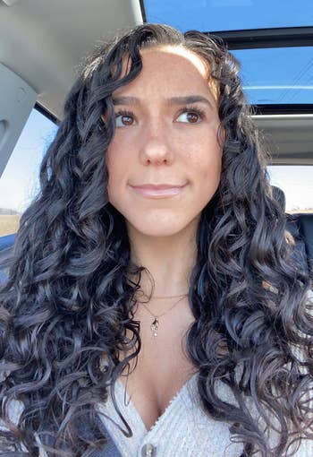 Person with long, curly hairsbreadth sits successful a car with a sunroof. They deterioration a textured apical with a delicate necklace, gazing upward