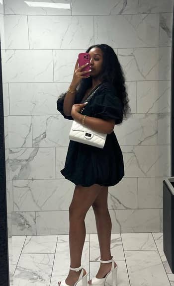 Person in black dress and white heels posing with a phone for a mirror selfie, holding a white handbag