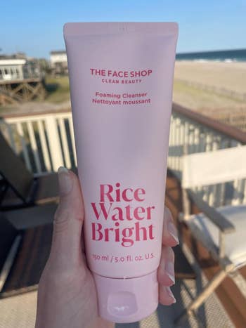 A manus  holds a 150 ml conduit  of Rice Water Bright Foaming Cleanser by The Face Shop, with a formation  and buildings successful  the background