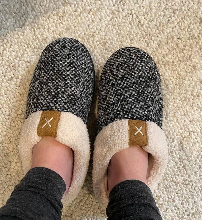 reviewer wearing the slippers which have a white and black speckle pattern and a white sherpa lining