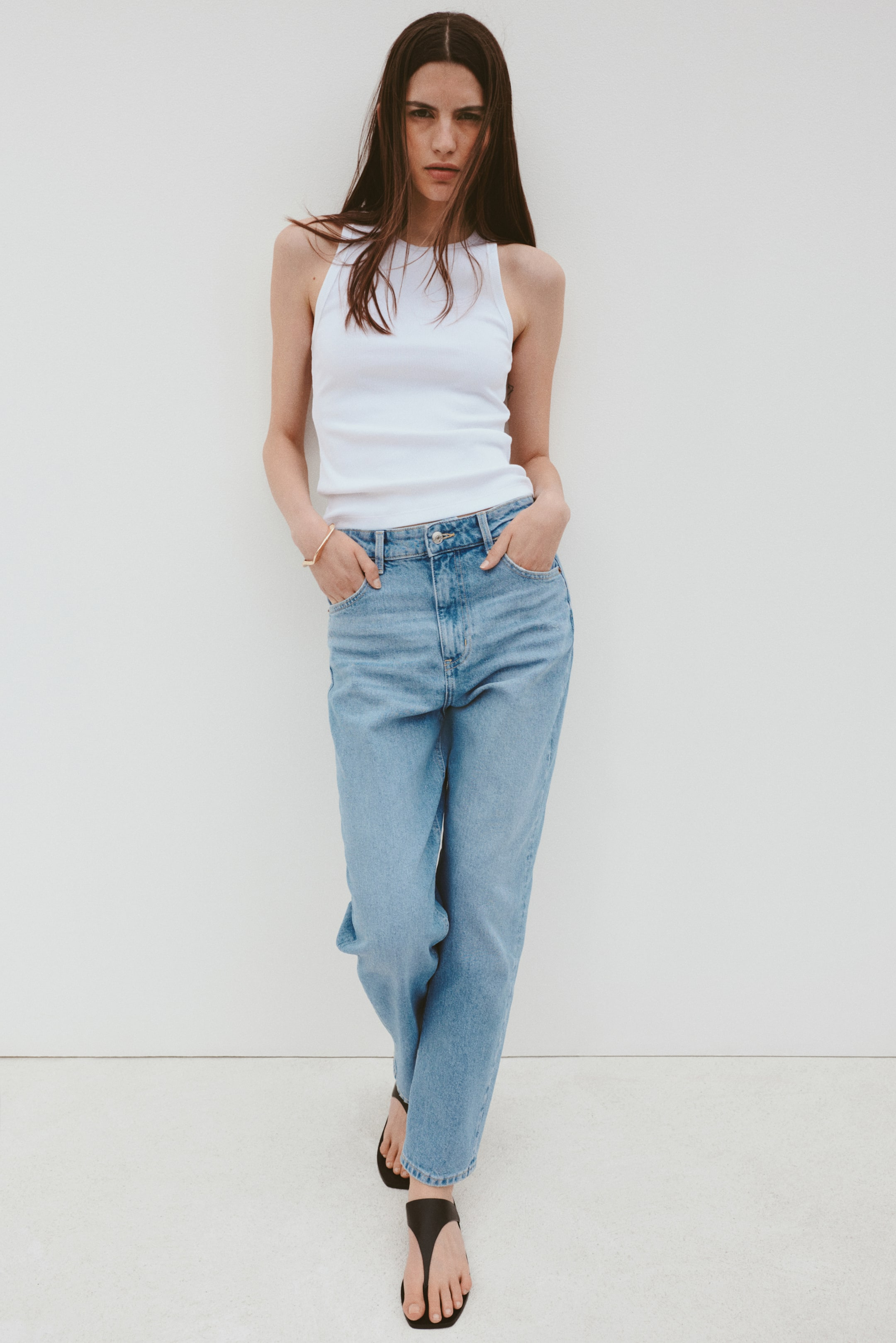 Person wearing a achromatic  vessel  top, relaxed-fit jeans, and sandals stands against a plain backdrop with hands successful  pockets, showcasing casual style