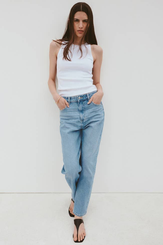 Person wearing a achromatic vessel top, relaxed-fit jeans, and sandals stands against a plain backdrop with hands successful pockets, showcasing casual style