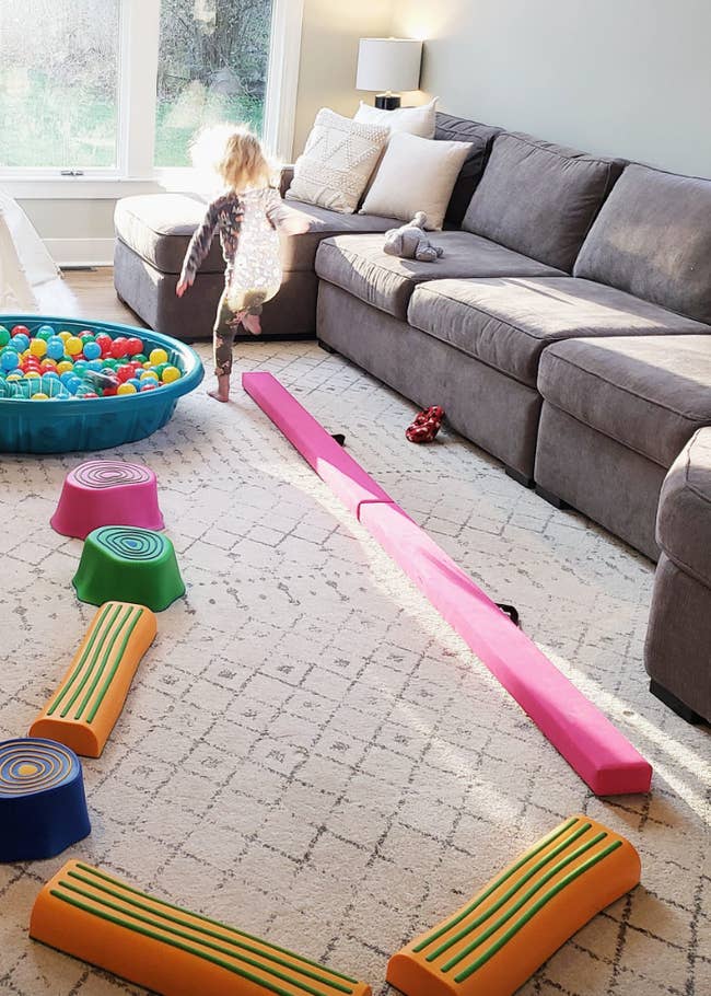 A child plays on a balance beam and stepping stones in a living room with a couch, ball pit, and large windows