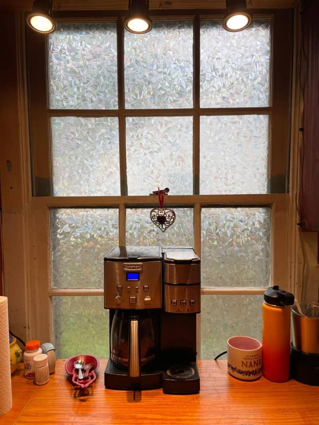 Kitchen counter with a coffee maker, mug, and vitamin bottles. Frosted glass window in the background with a heart decoration