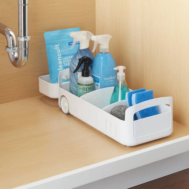 Cleaning supplies in a white caddy under a sink, including sprays, sponges, and detergent, suitable for organizing home cleaning essentials