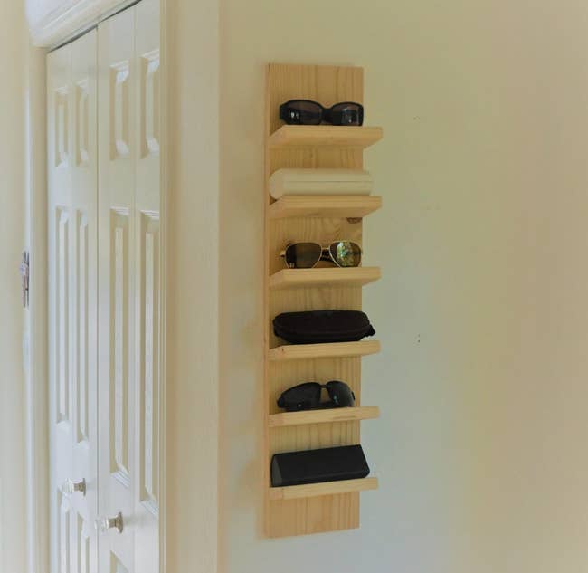 A natural wood colored organizer hanging on a wall with six sunglasses on it