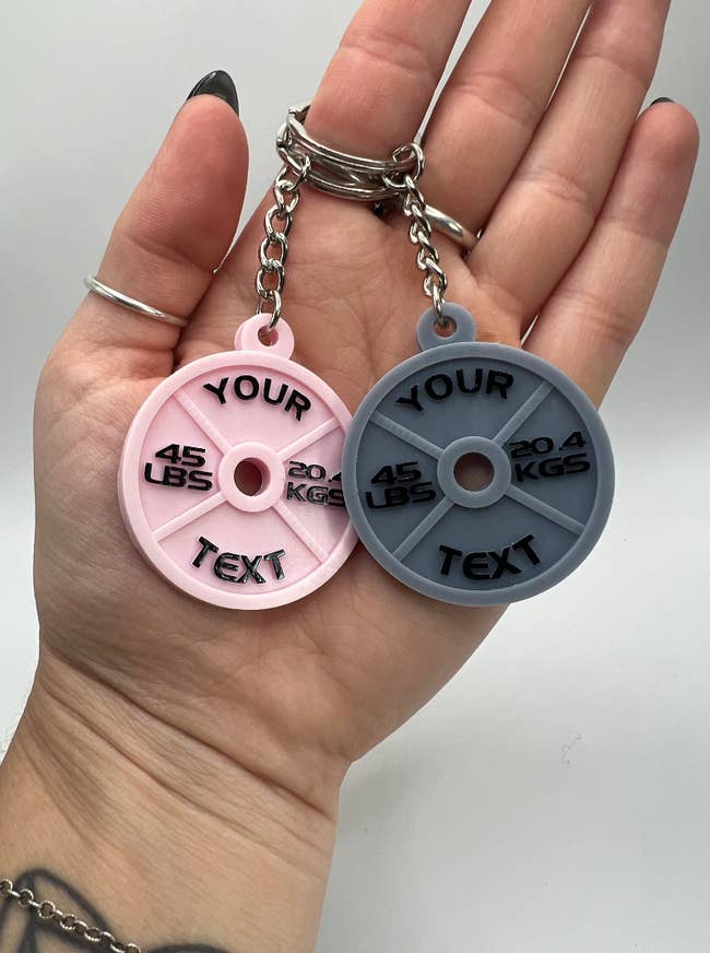 Model holding two weight-shaped keychains in pink and gray that have a weight and 
