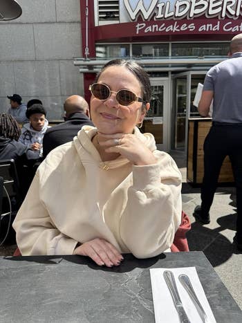 Person smiling successful  an outdoor café, wearing circular  sunglasses and a casual hoodie, with a paper   and cutlery connected  the table